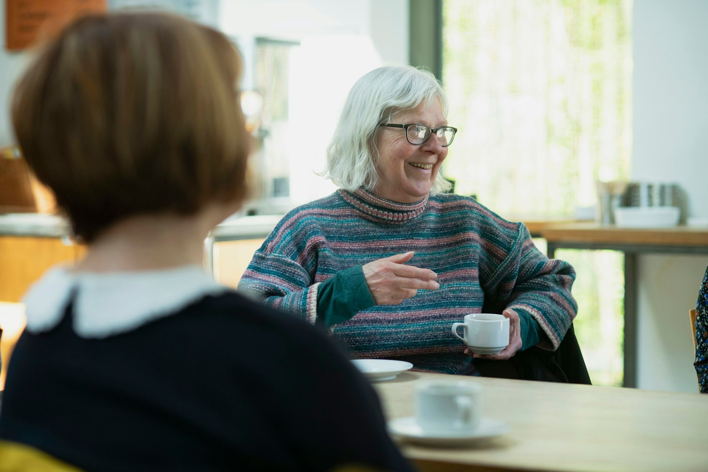 Zwei Frauen sitzen an einem Tisch und sprechen miteinander, vor sich Tassen